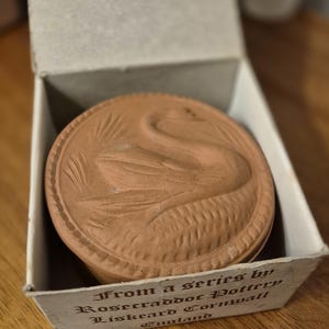 May include: A terracotta-coloured pottery piece with a swan design, in a cardboard box. The swan is embossed on the pottery. The box has text that reads "From a series by Rosecraddoc Pottery, Liskeard Cornwall, England."
