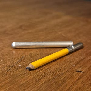 Puede incluir: Un lápiz amarillo con punta de metal plateado y tapa de metal plateado, sobre una superficie de madera. El lápiz tiene una punta de mina azul. Un tubo de metal plateado también está sobre la superficie de madera.