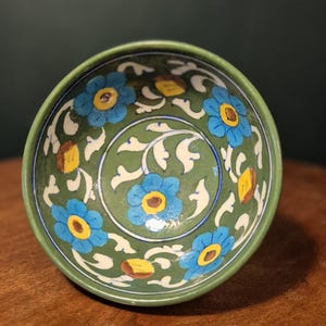Può includere: Una ciotola in ceramica con esterno e interno verdi. L'interno presenta un motivo floreale con fiori blu, centri gialli e accenti di foglie bianche. La ciotola è appoggiata su una superficie di legno.