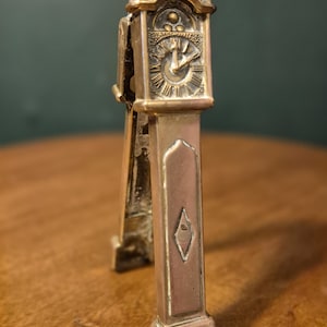 Könnte beinhalten: Ein miniaturförmiges Objekt in Form einer Uhr in Messingfarbe mit detailliertem Zifferblatt und dekorativen Akzenten. Das Objekt hat einen rechteckigen Körper mit einem rautenförmigen Design und einem verzierten Sockel. Die Uhr steht auf einer Holzoberfläche.