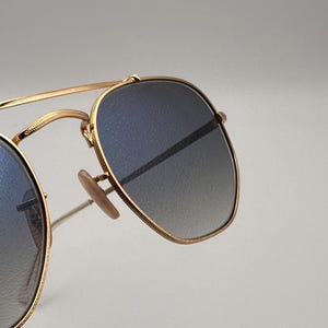May include: Gold-framed sunglasses with gradient blue lenses. The sunglasses feature a classic aviator design with a double bridge and nose pads. The lenses transition from a darker blue at the top to a lighter shade at the bottom. The frame is a shiny gold colour.