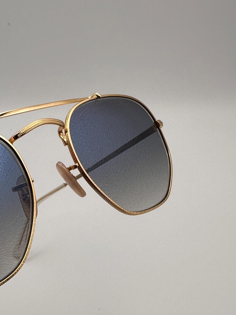 May include: Gold-framed sunglasses with gradient blue lenses. The sunglasses feature a classic aviator design with a double bridge and nose pads. The lenses transition from a darker blue at the top to a lighter shade at the bottom. The frame is a shiny gold colour.
