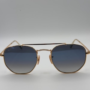 May include: Gold-coloured sunglasses with blue gradient lenses. The sunglasses feature a double bridge and a classic, geometric shape. The Ray-Ban logo is visible on the upper left lens. The design is sleek and stylish.