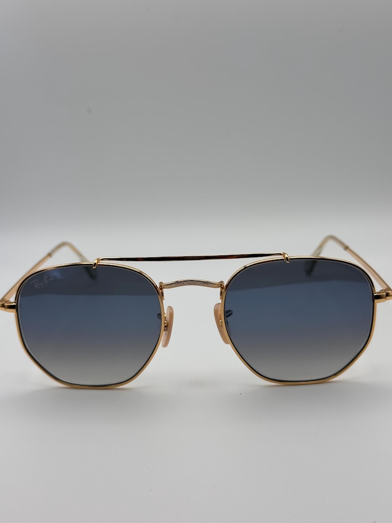 May include: Gold-coloured sunglasses with blue gradient lenses. The sunglasses feature a double bridge and a classic, geometric shape. The Ray-Ban logo is visible on the upper left lens. The design is sleek and stylish.