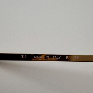 May include: Close-up of the temple of Ray-Ban sunglasses. The temple is a dark brown colour with the text "Ray-Ban" and "MADE IN ITALY" printed on it. The letter "R" and the letters "CE" are also visible.