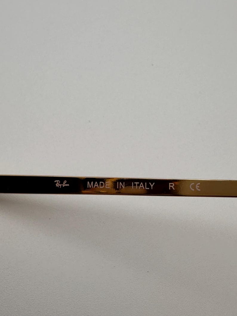 May include: Close-up of the temple of Ray-Ban sunglasses. The temple is a dark brown colour with the text "Ray-Ban" and "MADE IN ITALY" printed on it. The letter "R" and the letters "CE" are also visible.