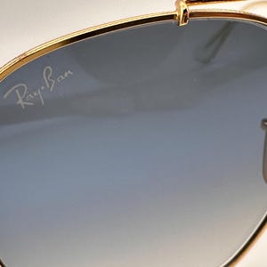 May include: Gold-coloured sunglasses with gradient blue lenses. The Ray-Ban logo is visible on the lens. The sunglasses have a classic aviator design.