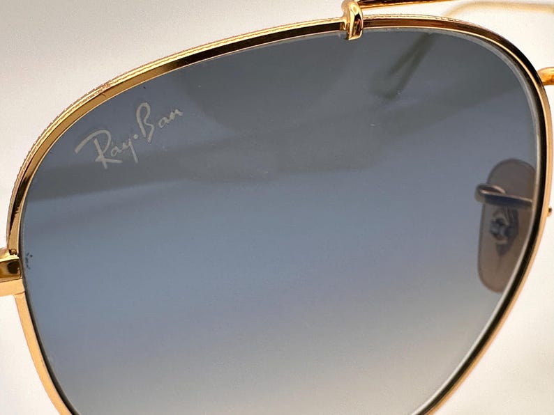 May include: Gold-coloured sunglasses with gradient blue lenses. The Ray-Ban logo is visible on the lens. The sunglasses have a classic aviator design.