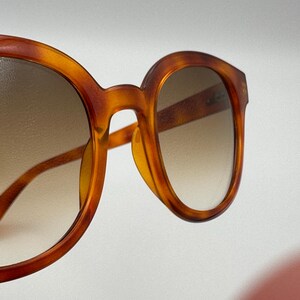 May include: Close-up of tortoiseshell sunglasses. The frames are a warm amber colour with a gradient lens. The round frames have a classic, vintage design, with a subtle nose bridge and temples.
