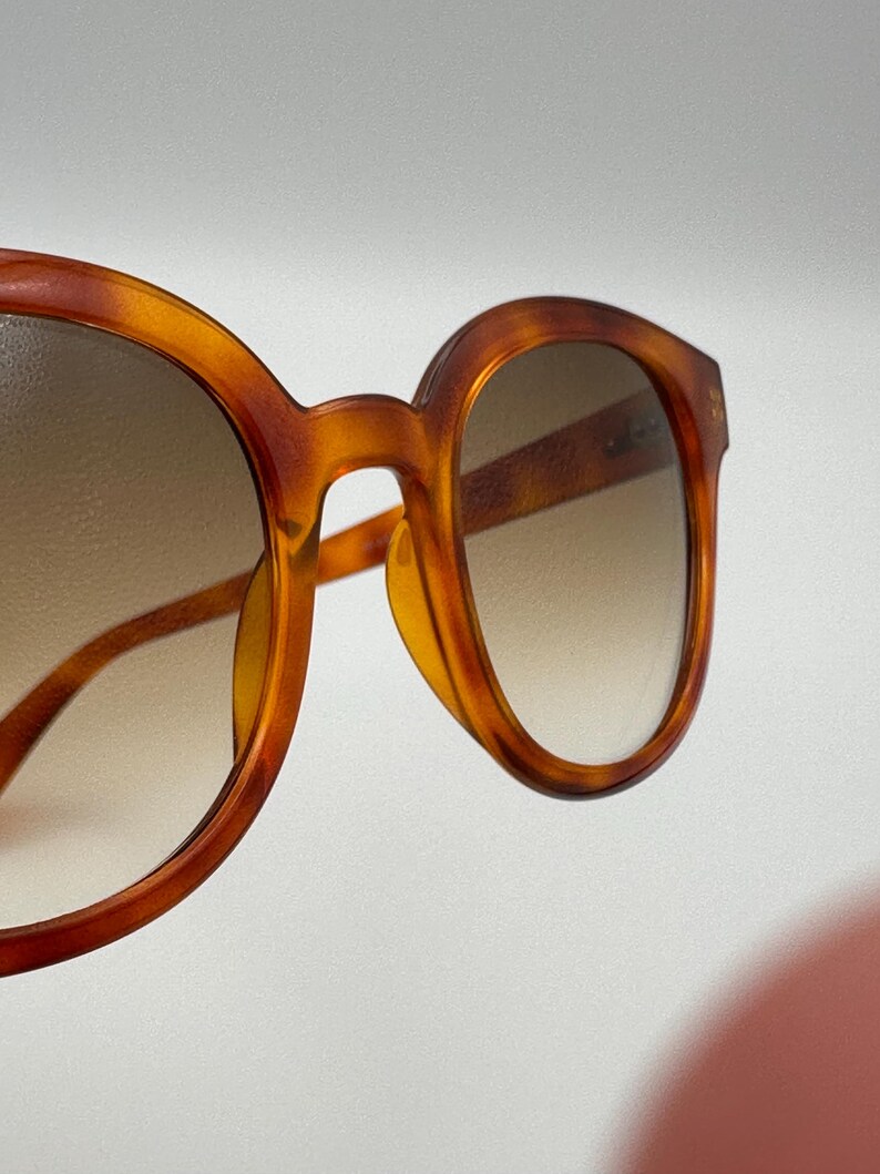 May include: Close-up of tortoiseshell sunglasses. The frames are a warm amber colour with a gradient lens. The round frames have a classic, vintage design, with a subtle nose bridge and temples.