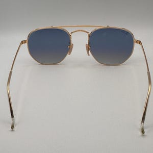 May include: Gold-coloured sunglasses with blue gradient lenses. The sunglasses have a double bridge and thin arms. The lenses are a hexagonal shape. The Ray-Ban logo is visible on the right lens.