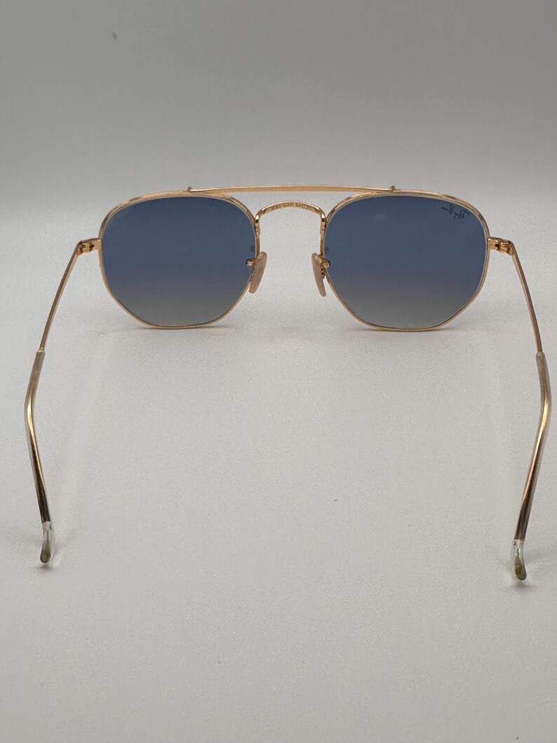 May include: Gold-coloured sunglasses with blue gradient lenses. The sunglasses have a double bridge and thin arms. The lenses are a hexagonal shape. The Ray-Ban logo is visible on the right lens.
