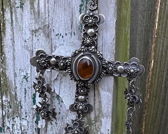 May include: A silver-toned cross pendant with intricate floral detailing and a central oval amber-colored stone. The cross hangs from a brown cord and features dangling floral accents. The background is a weathered wooden surface.
