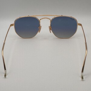 May include: Gold-coloured sunglasses with blue gradient lenses. The sunglasses have a double bridge and an aviator shape. The temples are thin and extend to the ear pieces. The lenses are a cool blue colour.