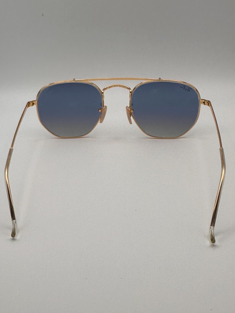 May include: Gold-coloured sunglasses with blue gradient lenses. The sunglasses have a double bridge and an aviator shape. The temples are thin and extend to the ear pieces. The lenses are a cool blue colour.