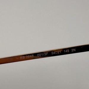May include: Close-up of the arm of gold-coloured eyeglasses. The text "RB 3648 001/3F 54021 145 2N" is printed on the arm. The eyeglasses are likely for vision correction or sun protection.