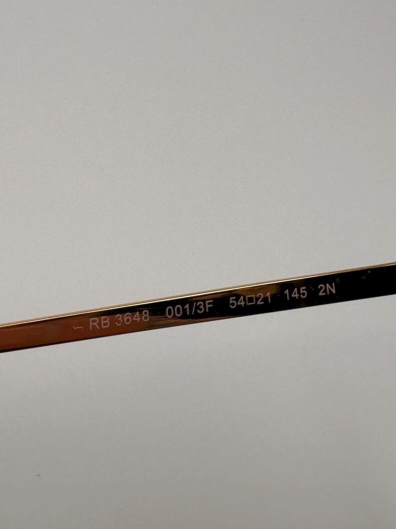 May include: Close-up of the arm of gold-coloured eyeglasses. The text "RB 3648 001/3F 54021 145 2N" is printed on the arm. The eyeglasses are likely for vision correction or sun protection.