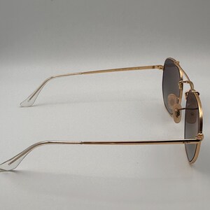 May include: Gold-coloured sunglasses with dark lenses. The sunglasses have a double bridge and a classic aviator shape. The temples are thin and gold, with clear plastic earpieces. The lenses are a gradient shade of brown.