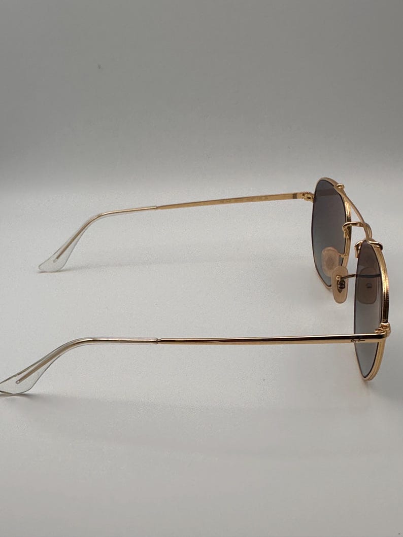 May include: Gold-coloured sunglasses with dark lenses. The sunglasses have a double bridge and a classic aviator shape. The temples are thin and gold, with clear plastic earpieces. The lenses are a gradient shade of brown.