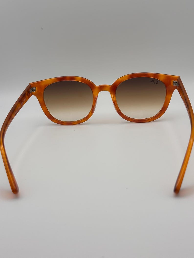 May include: Amber tortoiseshell sunglasses with brown gradient lenses. The frames have a classic round shape. The arms extend outwards, showcasing the full design. The sunglasses are set against a plain white background. The lenses are a brown gradient.