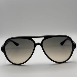 Puede incluir: Gafas de sol aviador negras con lentes degradadas. Las gafas tienen una montura negra gruesa y brillante y una forma cl&aacute;sica de l&aacute;grima. Las lentes pasan de un gris oscuro en la parte superior a un tono m&aacute;s claro en la parte inferior.