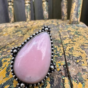 May include: A large, teardrop-shaped ring featuring a pale pink gemstone set in a silver-toned metal. The ring's bezel is adorned with small, round beads. The ring is displayed on a distressed, yellow and green wooden surface.