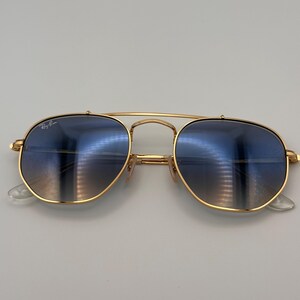 May include: Gold-coloured sunglasses with blue gradient lenses. The sunglasses feature a double bridge and clear nose pads. The Ray-Ban logo is visible on the upper left lens. The lenses reflect the surrounding environment.