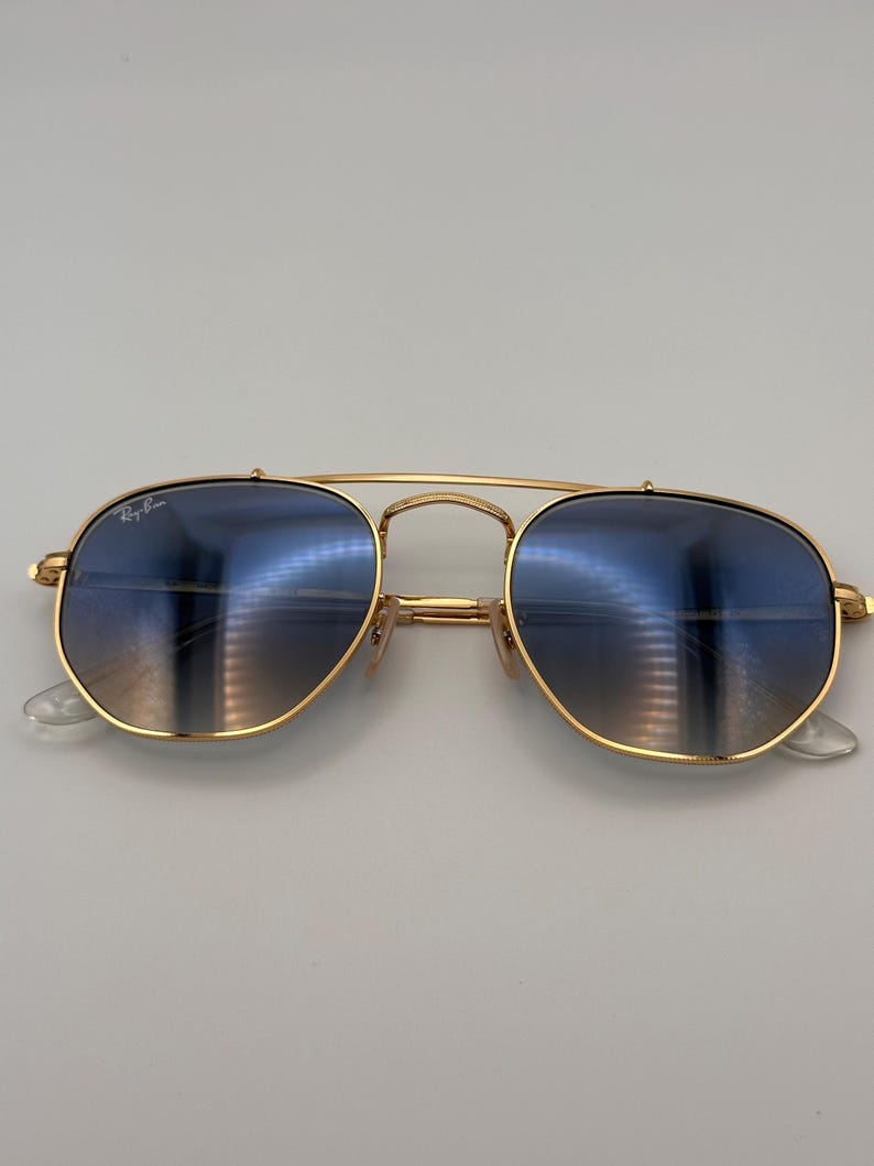 May include: Gold-coloured sunglasses with blue gradient lenses. The sunglasses feature a double bridge and clear nose pads. The Ray-Ban logo is visible on the upper left lens. The lenses reflect the surrounding environment.