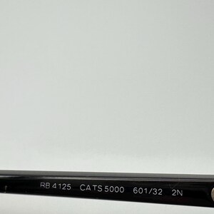 Puede incluir: Primer plano de la patilla de unas gafas de sol negras. El texto "RB 4125 CATS 5000 601/32 2N" est&aacute; impreso en blanco en la patilla. El fondo es gris claro.