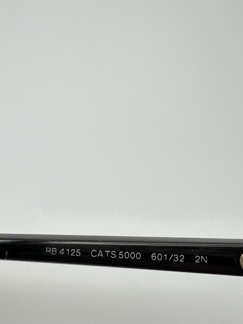 Puede incluir: Primer plano de la patilla de unas gafas de sol negras. El texto "RB 4125 CATS 5000 601/32 2N" est&aacute; impreso en blanco en la patilla. El fondo es gris claro.