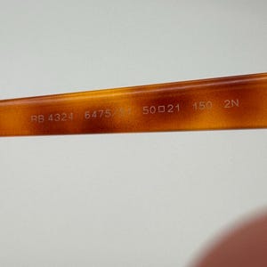 May include: Close-up of the inner arm of tortoiseshell eyeglasses. The text "RB 4324 6475/31 50 21 150 2N" is printed in white on the brown frame. The frame is a warm, amber colour.