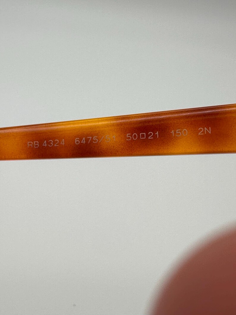 May include: Close-up of the inner arm of tortoiseshell eyeglasses. The text "RB 4324 6475/31 50 21 150 2N" is printed in white on the brown frame. The frame is a warm, amber colour.
