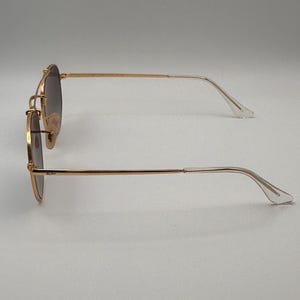 May include: Gold-coloured sunglasses with gradient lenses. The glasses have a classic round shape with a double bridge and thin arms. The lenses transition from dark at the top to lighter at the bottom. The arms curve gently to the ear pieces.