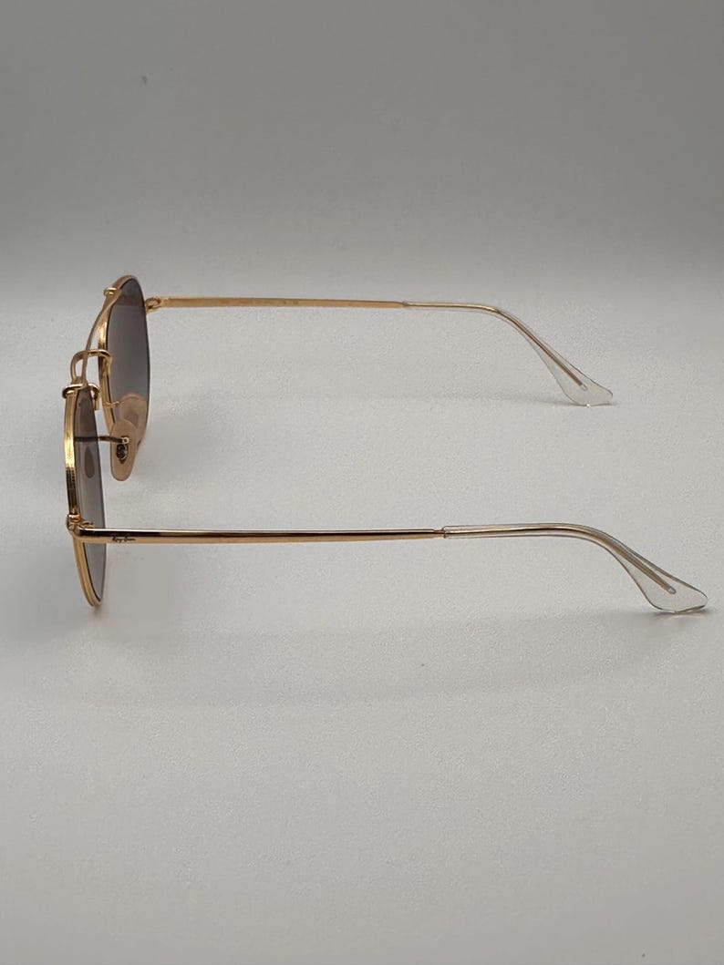 May include: Gold-coloured sunglasses with gradient lenses. The glasses have a classic round shape with a double bridge and thin arms. The lenses transition from dark at the top to lighter at the bottom. The arms curve gently to the ear pieces.