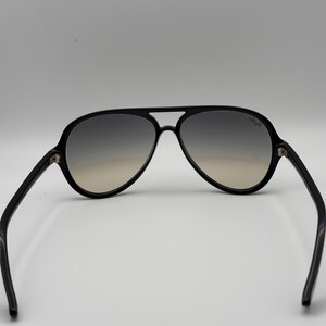 Puede incluir: Gafas de sol aviador negras con lentes degradadas. Las gafas tienen una montura y patillas negras gruesas. Las lentes pasan de gris oscuro en la parte superior a marr&oacute;n claro en la inferior. El logotipo de Ray-Ban es visible en la lente derecha.