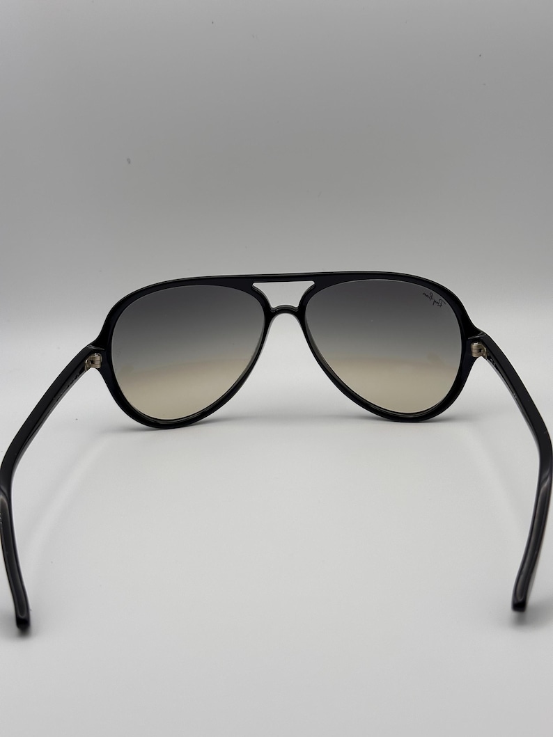 Puede incluir: Gafas de sol aviador negras con lentes degradadas. Las gafas tienen una montura y patillas negras gruesas. Las lentes pasan de gris oscuro en la parte superior a marr&oacute;n claro en la inferior. El logotipo de Ray-Ban es visible en la lente derecha.