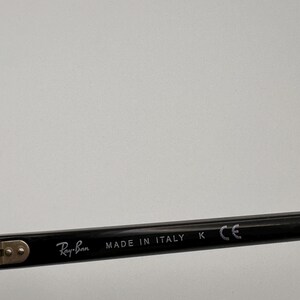 Puede incluir: Primer plano de una patilla de gafas Ray-Ban negra. El texto "Ray-Ban" est&aacute; impreso en blanco, seguido de "MADE IN ITALY", "K" y "CE". La patilla est&aacute; sobre un fondo liso y claro.