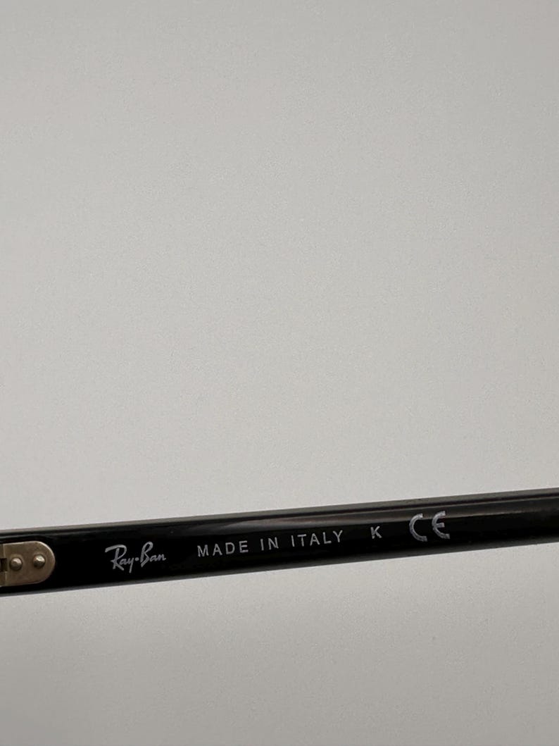 Puede incluir: Primer plano de una patilla de gafas Ray-Ban negra. El texto "Ray-Ban" est&aacute; impreso en blanco, seguido de "MADE IN ITALY", "K" y "CE". La patilla est&aacute; sobre un fondo liso y claro.