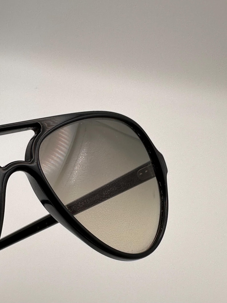 Puede incluir: Gafas de sol aviador negras con doble puente y lentes degradadas. Las gafas tienen una montura gruesa y un dise&ntilde;o cl&aacute;sico. Las lentes pasan de un tono m&aacute;s oscuro en la parte superior a un tono m&aacute;s claro en la parte inferior.