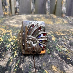May include: A silver-toned ring featuring a Native American chief design. The ring's face showcases a detailed portrait with a feathered headdress in red, white, and blue. The ring is set against a weathered wooden surface.
