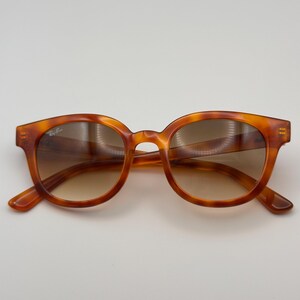 May include: Tortoiseshell sunglasses with a rounded frame and gradient lenses. The sunglasses have a classic design. The Ray-Ban logo is visible on the left lens. Suitable for everyday wear.