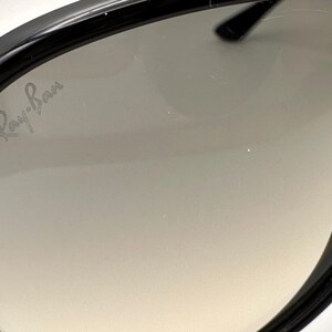 Puede incluir: Primer plano de gafas de sol negras con lentes degradadas. El logotipo de Ray-Ban es visible en la lente. Las gafas de sol tienen un dise&ntilde;o elegante y moderno con un acabado brillante. Las lentes hacen una transici&oacute;n de un tono m&aacute;s oscuro en la parte superior a un tono m&aacute;s claro en la parte inferior.