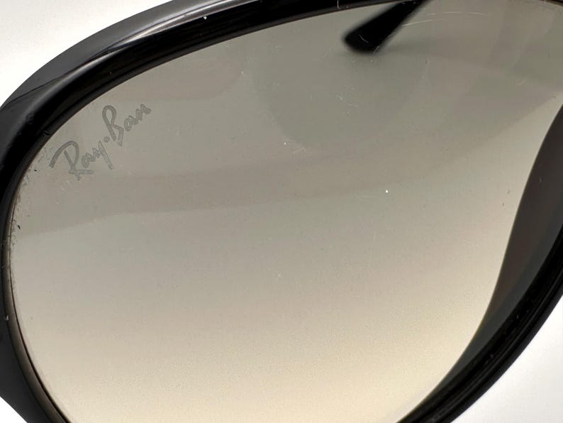 Puede incluir: Primer plano de gafas de sol negras con lentes degradadas. El logotipo de Ray-Ban es visible en la lente. Las gafas de sol tienen un dise&ntilde;o elegante y moderno con un acabado brillante. Las lentes hacen una transici&oacute;n de un tono m&aacute;s oscuro en la parte superior a un tono m&aacute;s claro en la parte inferior.