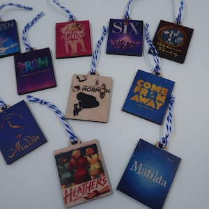 May include: Ten wooden ornaments featuring Broadway show posters. The ornaments are square and have blue and white twine attached. The shows featured are Frozen, Mean Girls, Six, The Addams Family, The Prom, The Book of Mormon, Come From Away, Aladdin, Heathers, and Matilda.