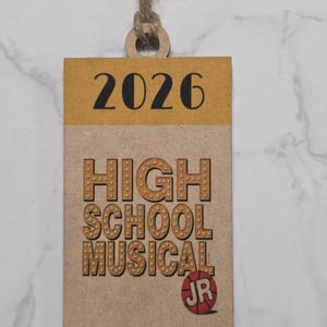 May include: A rectangular wooden tag with the text "2026" in black at the top, and "HIGH SCHOOL MUSICAL JR" in brown, with a basketball graphic. The tag is attached to a natural twine loop.