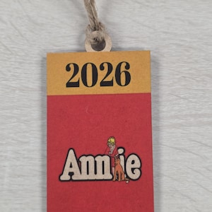 May include: A rectangular tag with a jute string for hanging. The top section is yellow with the black text "2026". The bottom section is red with the word "Annie" in a retro font and an illustration of a girl and dog.