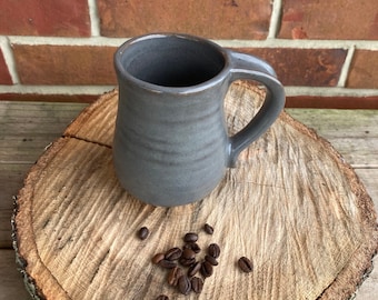 May include: A gray ceramic coffee mug with a curved handle sits on a wooden slice, with coffee beans scattered nearby. The mug has a simple, classic design and a smooth, matte finish. The background is a brick wall.