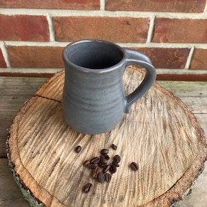 May include: A gray ceramic coffee mug with a curved handle sits on a wooden slice, with coffee beans scattered nearby. The mug has a simple, classic design and a smooth, matte finish. The background is a brick wall.
