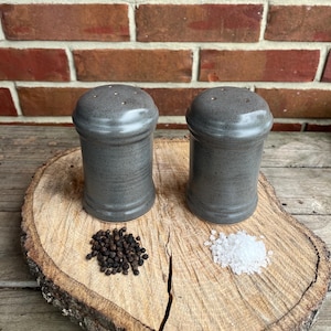 May include: Two gray ceramic salt and pepper shakers on a wooden surface. The shakers have a textured finish and are filled with salt and pepper.