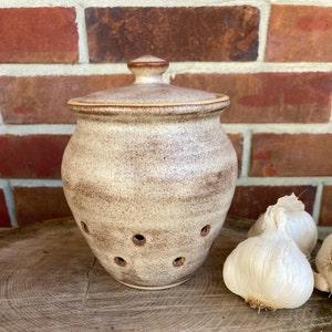 May include: A white ceramic garlic keeper with brown speckles and holes for ventilation. The keeper has a lid and is sitting on a wooden surface with two whole garlic bulbs.
